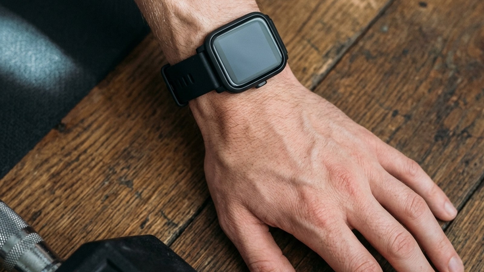 A close-up overhead view of a wrist wearing a generic black sports smartwatch with a completely blank dark display, hand resting palm-down on a wooden gym floor.